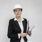 Confident female architect in a suit and hard hat holding blueprints against a white background.